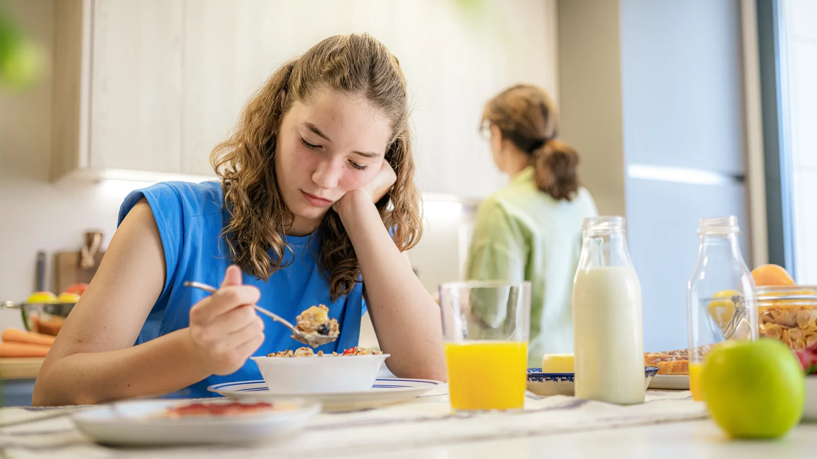 Teen girl sitting at breakfast while a parent stands nearby for an anorexia red flags guide